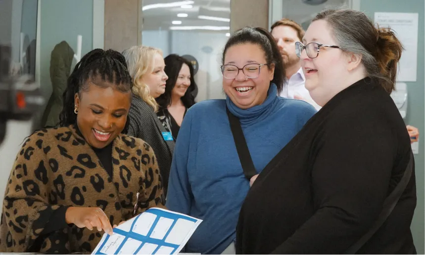 Three Alberta Blue Cross employees laughing and talking while looking at a printed paper during an office event, with colleagues chatting in the background.