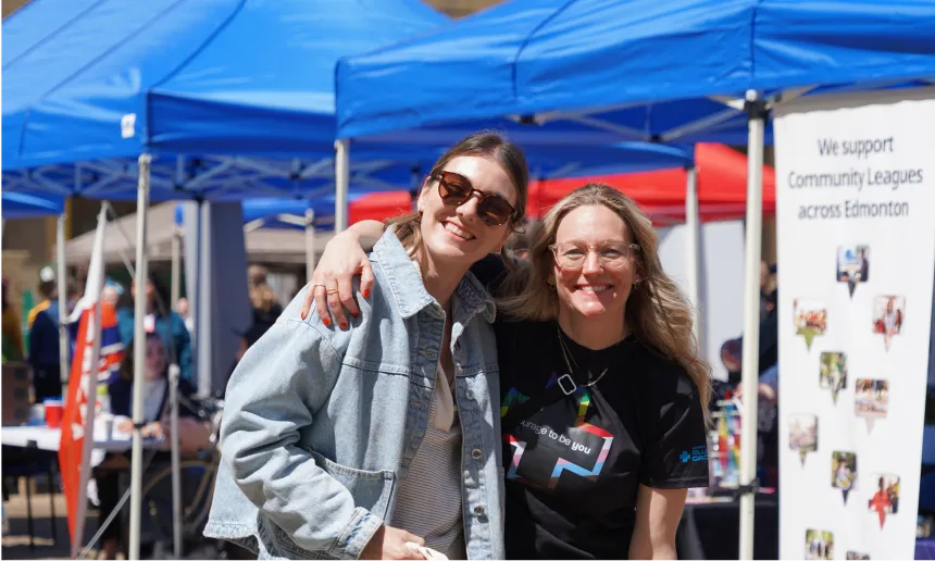 Two Alberta Blue Cross employees smiling and standing together at an outdoor event, with blue and red tents in the background.